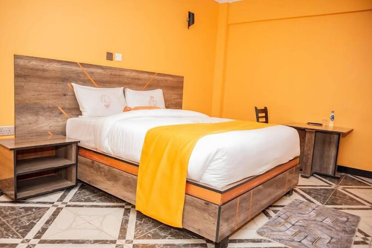 Una habitación de hotel con estructura de cama de madera, ropa de cama blanca, una manta amarilla y una mesita de noche con lámpara.