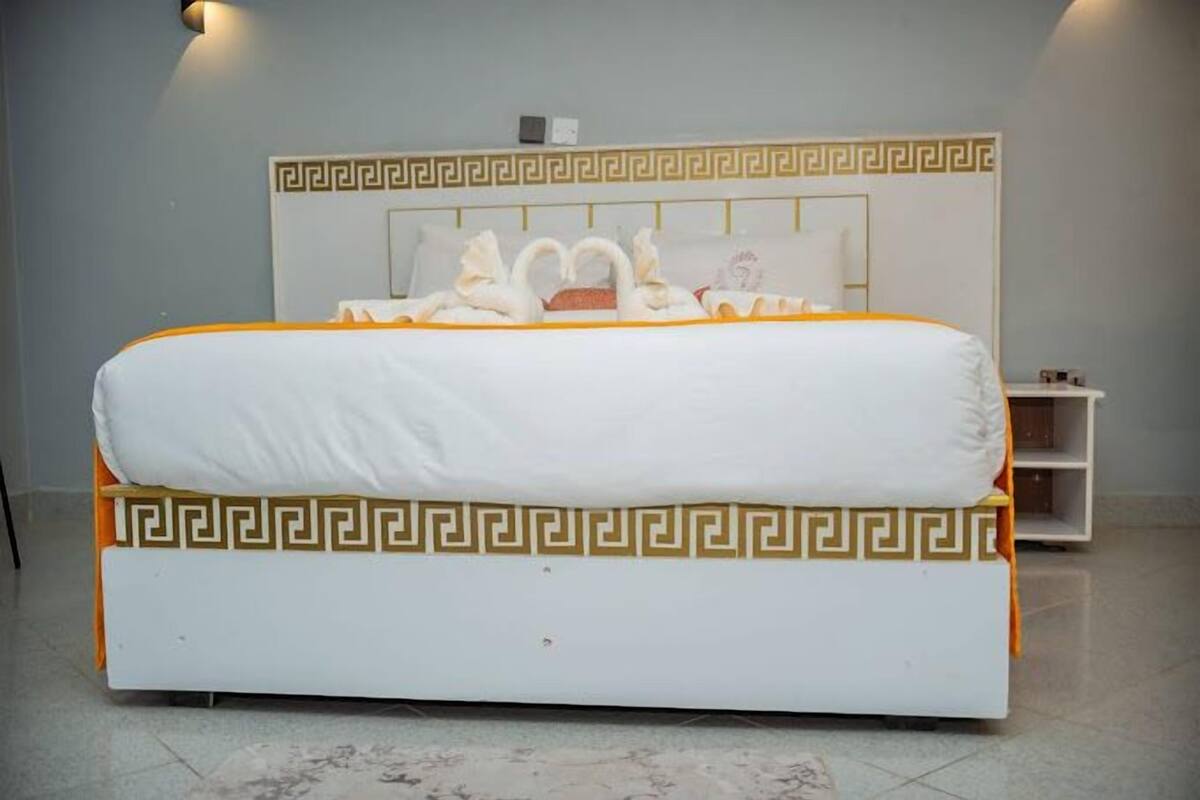 Una cama con cabecera blanca y dorada, almohadas con forma de cisne y colcha blanca.