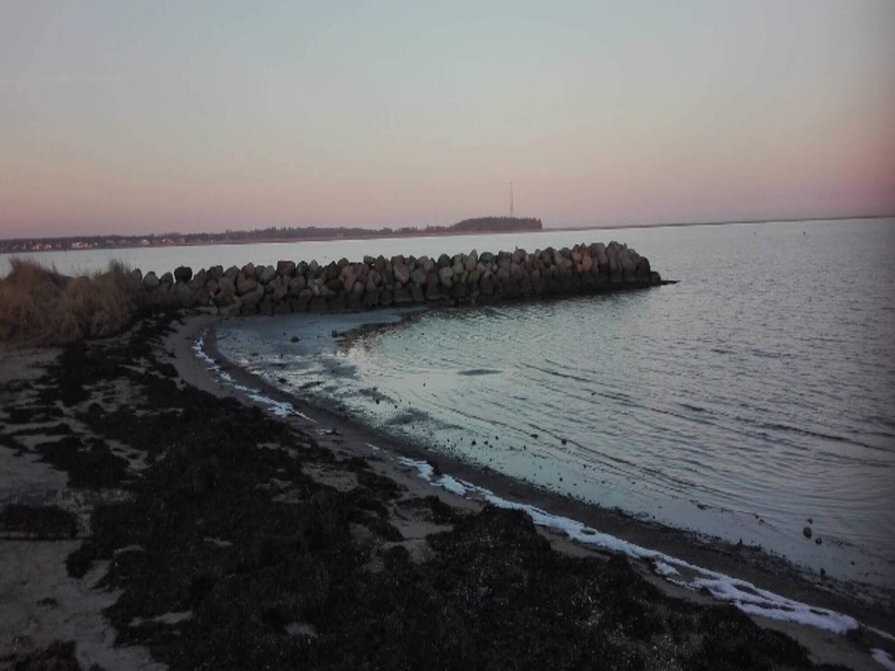 A coastal scene with a stone breakwater extending into calm waters, a sandy beach, and a distant shoreline with buildings.
