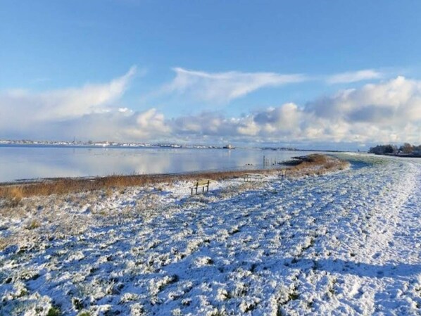Schneebedeckte Landschaft mit einer zugefrorenen Wasserfläche, blauem Himmel und Gebäuden in der Ferne.