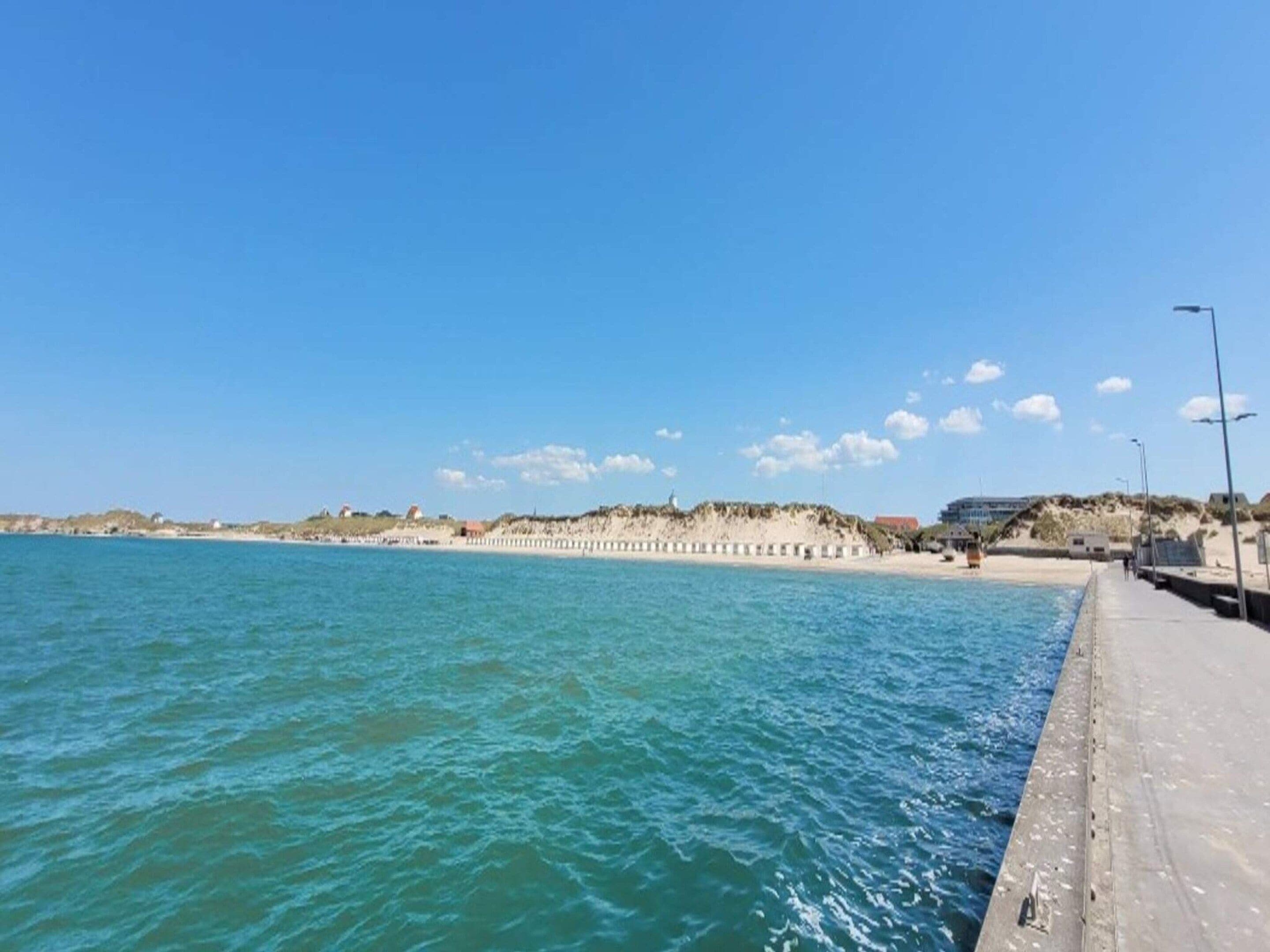 Escena costera con un paseo marítimo de hormigón a lo largo del agua, una playa arenosa y una colina distante con edificios.
