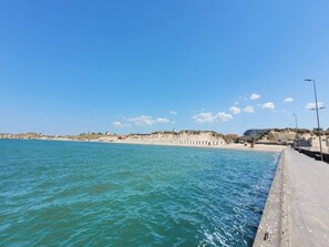 Eine Küstenlandschaft mit einem Betonsteg entlang des Wassers, einem Sandstrand und einer entfernten Anhöhe mit Gebäuden.