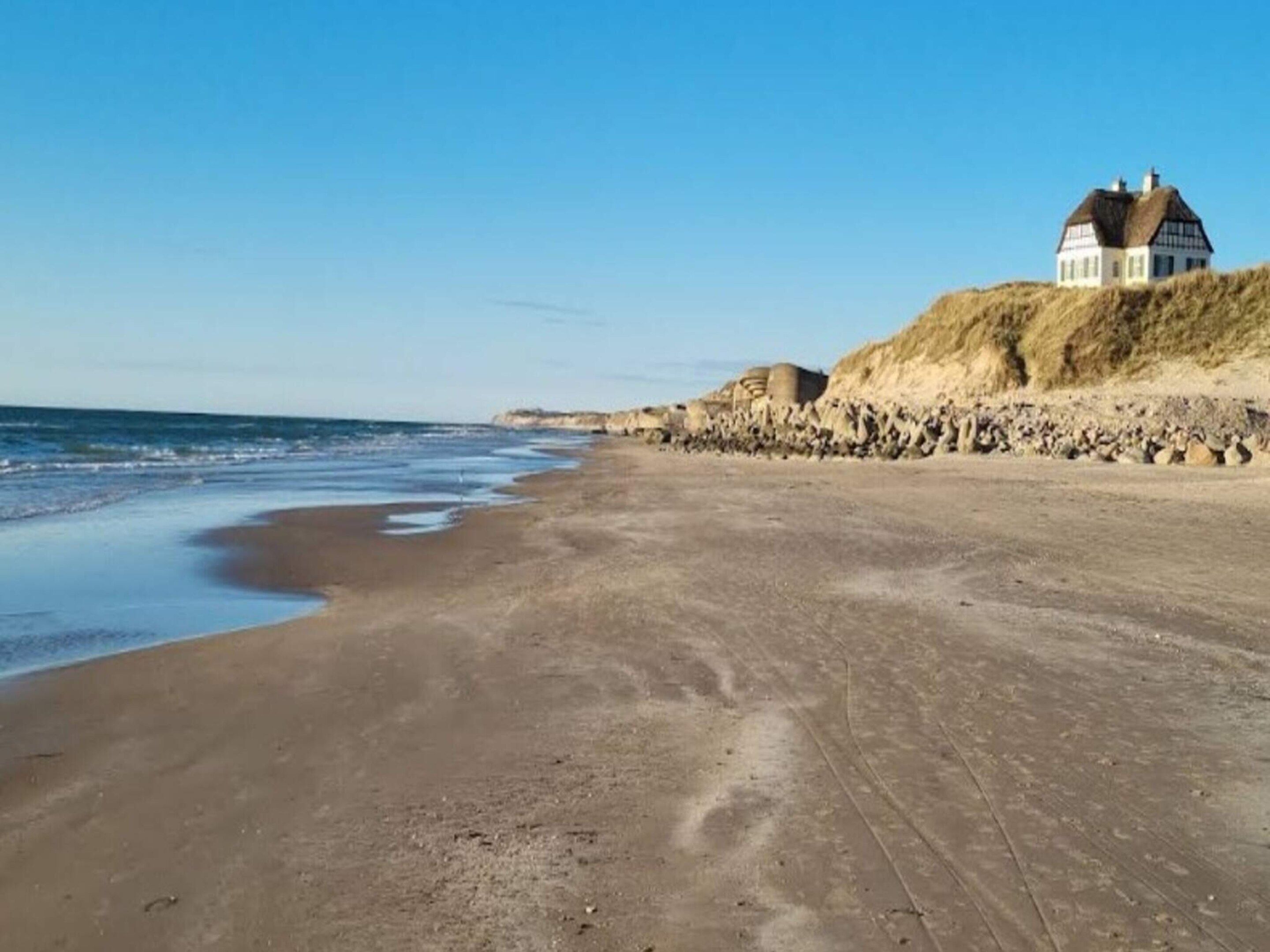A beach with a house on a dune, clear skies, and calm sea.