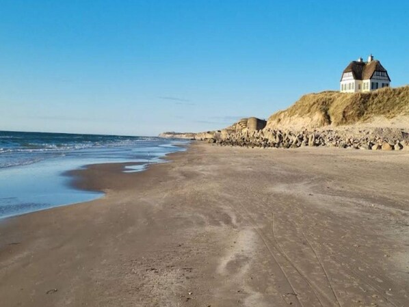 A beach with a house on a dune, clear skies, and calm sea. - 7 Person Holiday Home in Vaeggerlose (Vaeggerlose)