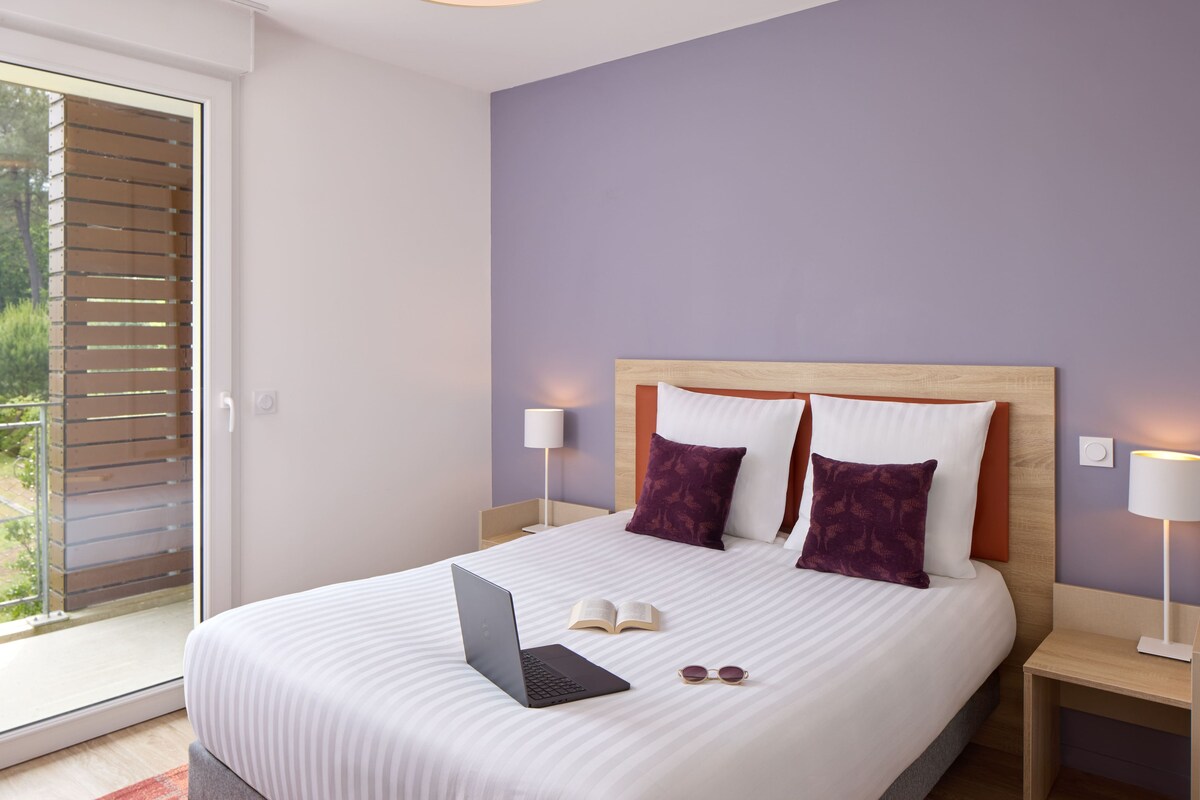 A neatly made bed with a laptop, book, and sunglasses on it, a wooden headboard, and two bedside lamps.