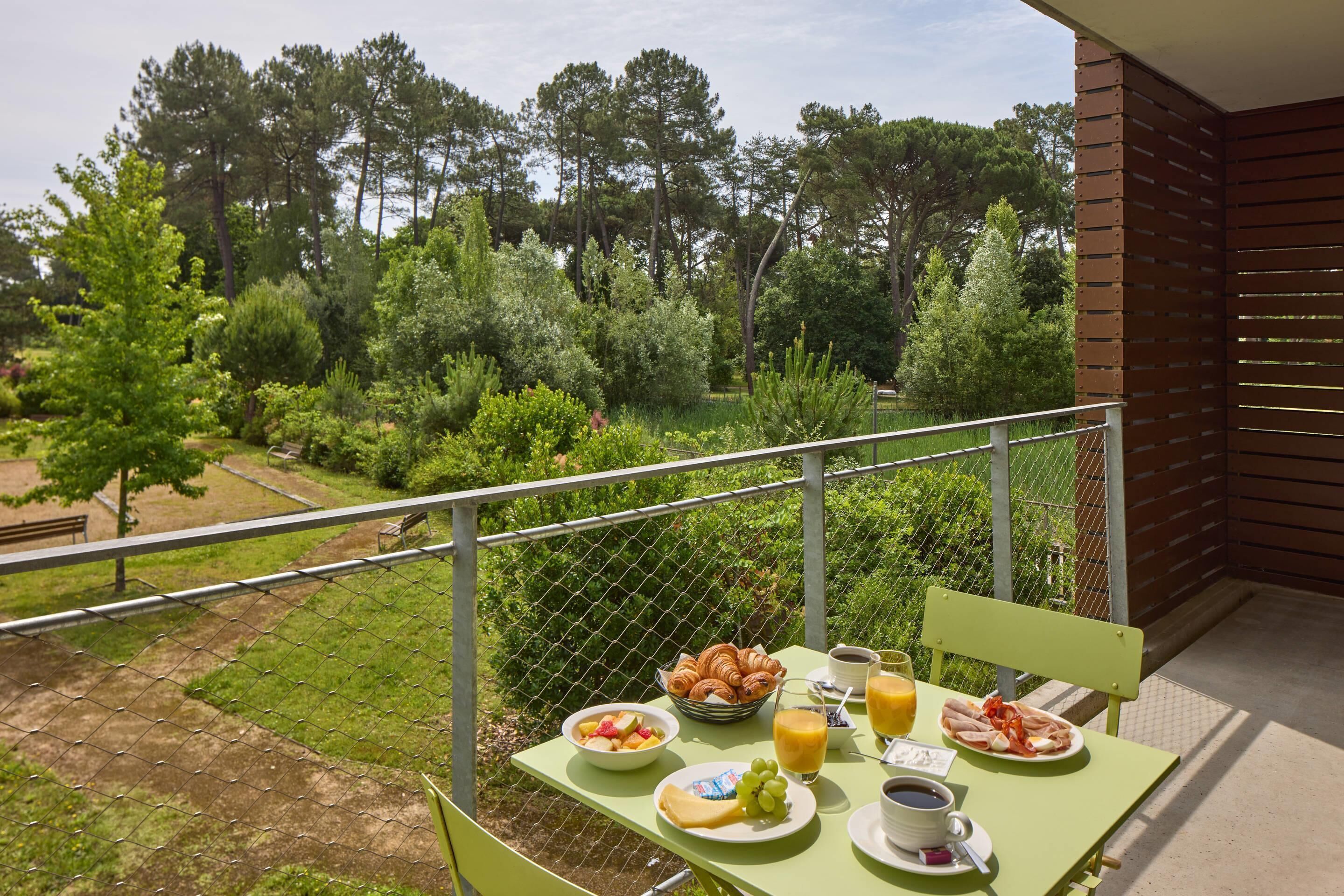A balcony with breakfast items including croissants, fruit, and coffee, overlooking a lush garden and trees.