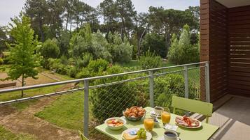 A balcony with breakfast items including croissants, fruit, and coffee, overlooking a lush garden and trees.