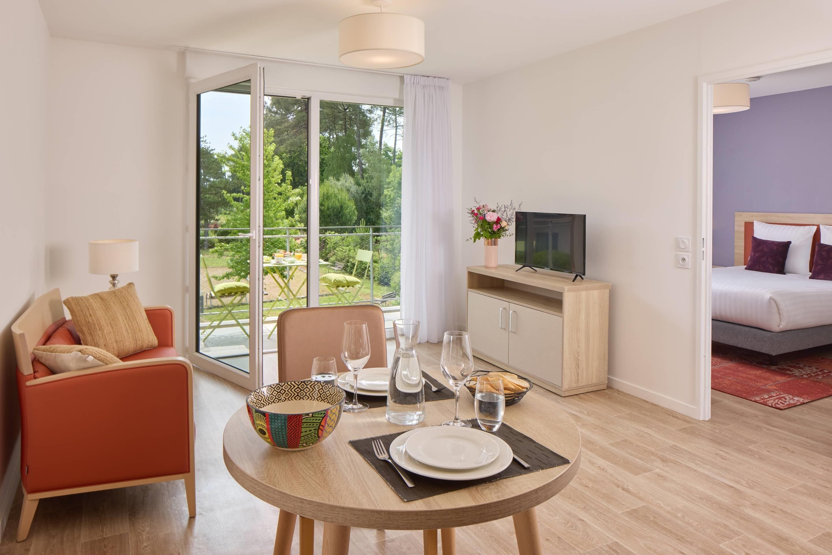 Een moderne hotelkamer met een eethoek, een televisie en een balkon met uitzicht op groen.