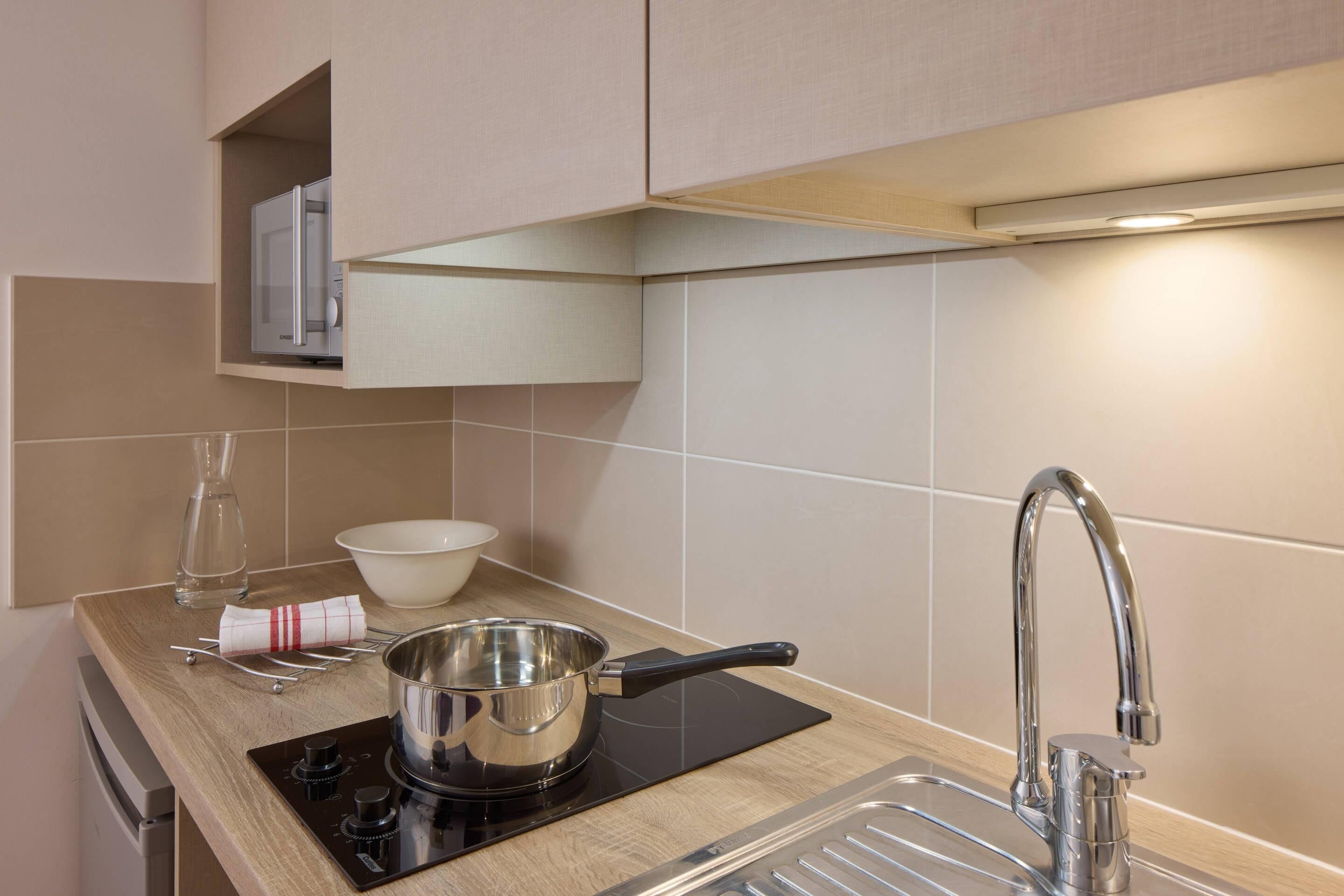 Et moderne køkken med indbygget mikrobølgeovn, en gryde i rustfrit stål på en induktionskogeplade, en vask med moderne armatur og en glasvase på bordpladen.
