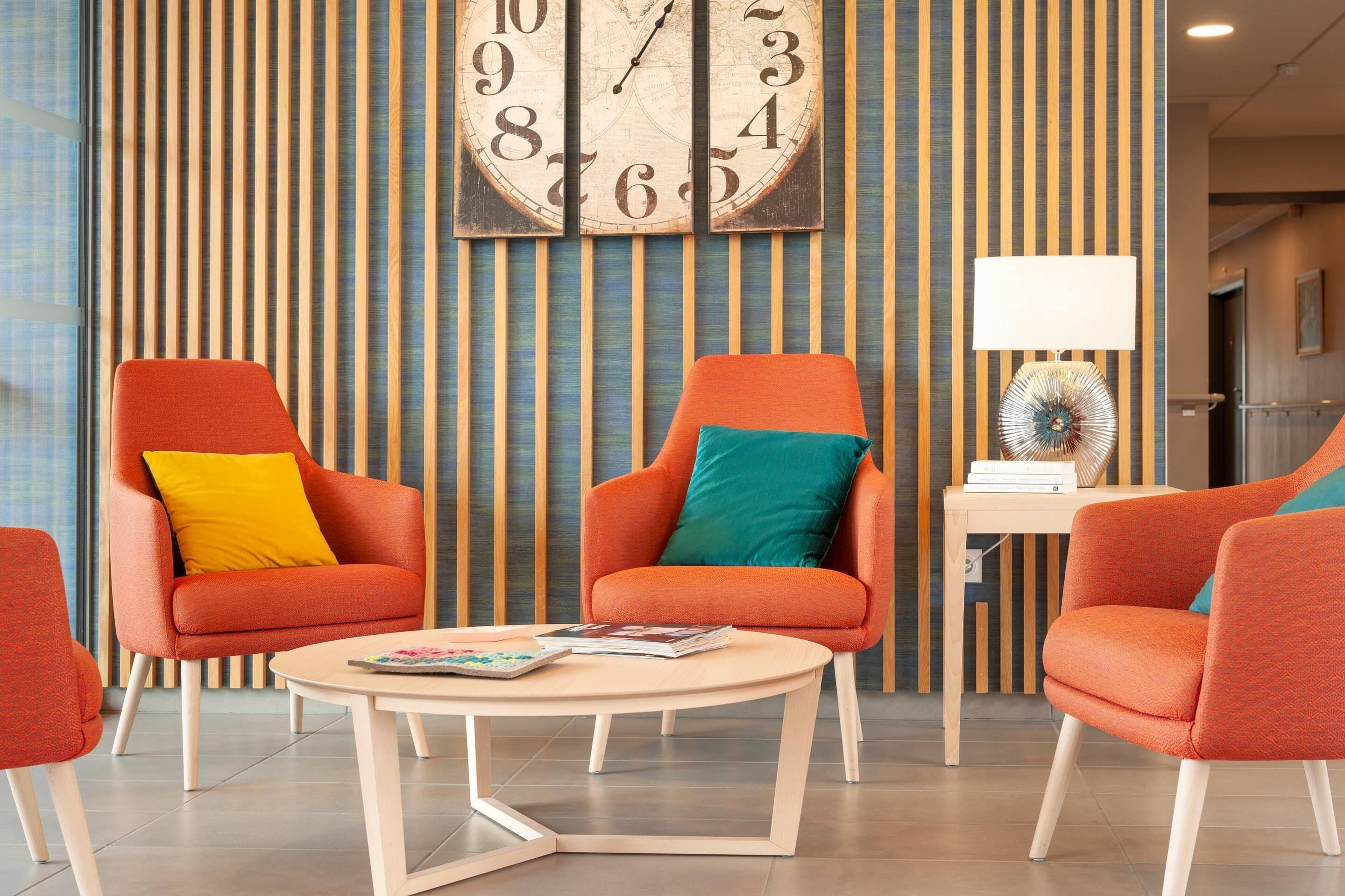 Eine moderne Hotelhalle mit Streifenmuster-Tapete, zwei orangefarbene Sessel mit bunten Kissen, ein runder Holztisch und eine große Wanduhr.