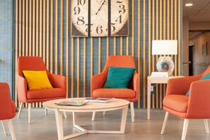 Un hall d’hôtel moderne avec un papier peint à rayures, deux fauteuils orange ornés de coussins colorés, une table ronde en bois et une grande horloge murale.