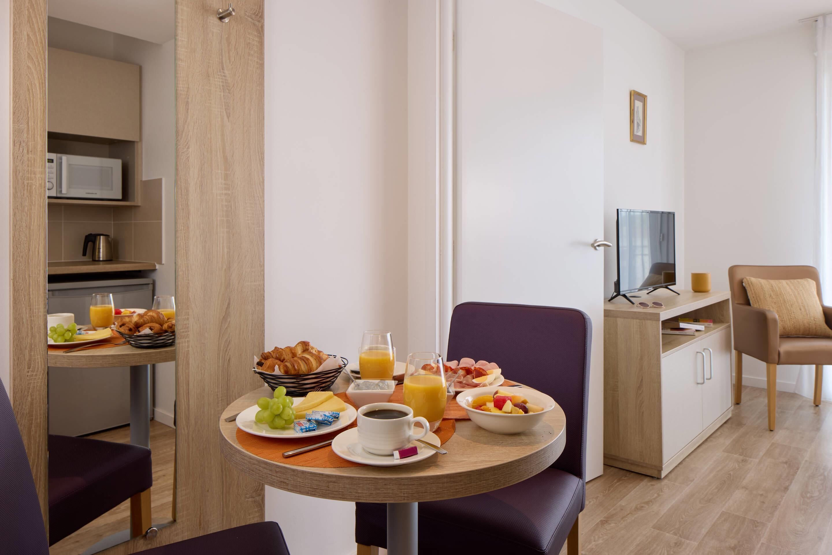 A modern hotel room with a dining area featuring breakfast items, a television, and a comfortable seating arrangement.