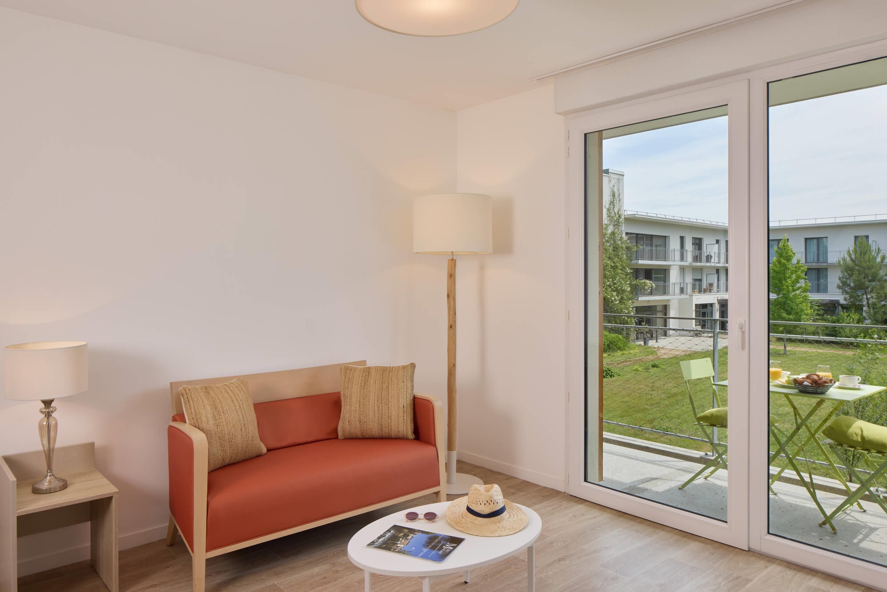 Uma sala de estar moderna com um sofá vermelho, um abajur e vista para uma varanda com mobiliário exterior.
