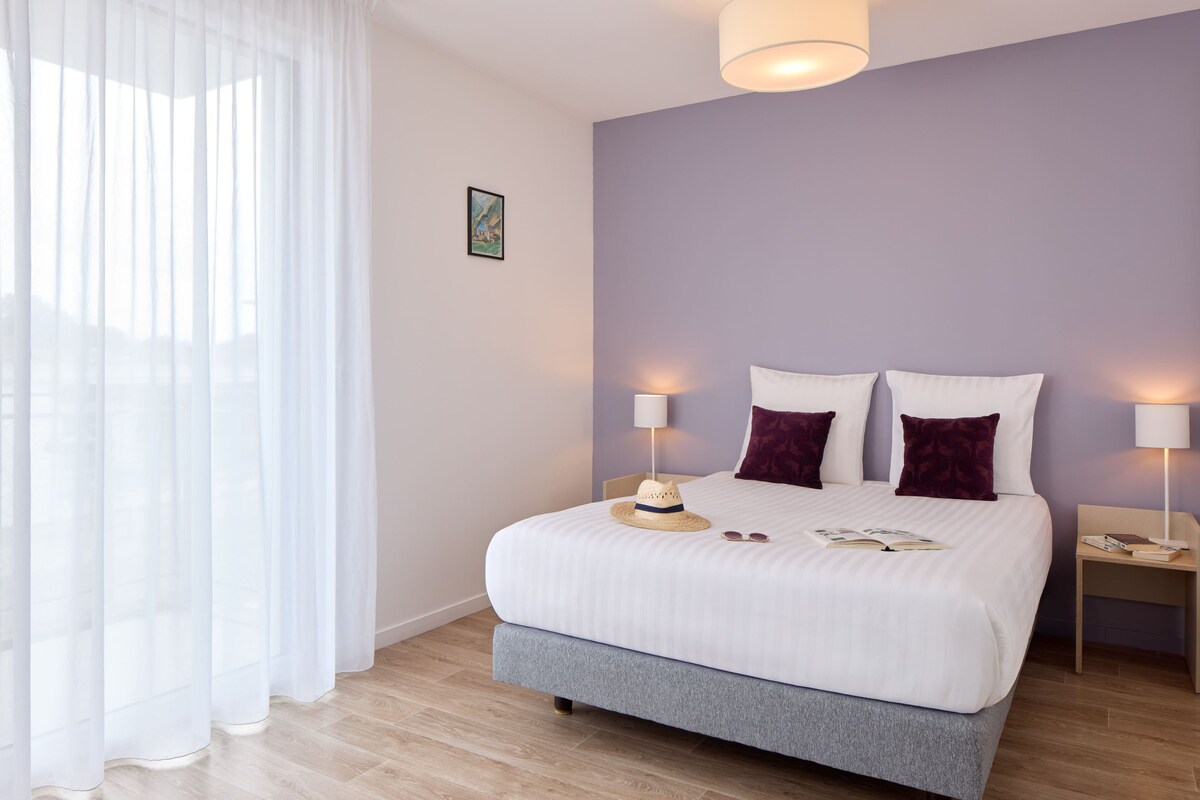 A neatly made bed with white bedding and two pillows, a bedside table with a lamp, and a window with sheer curtains.
