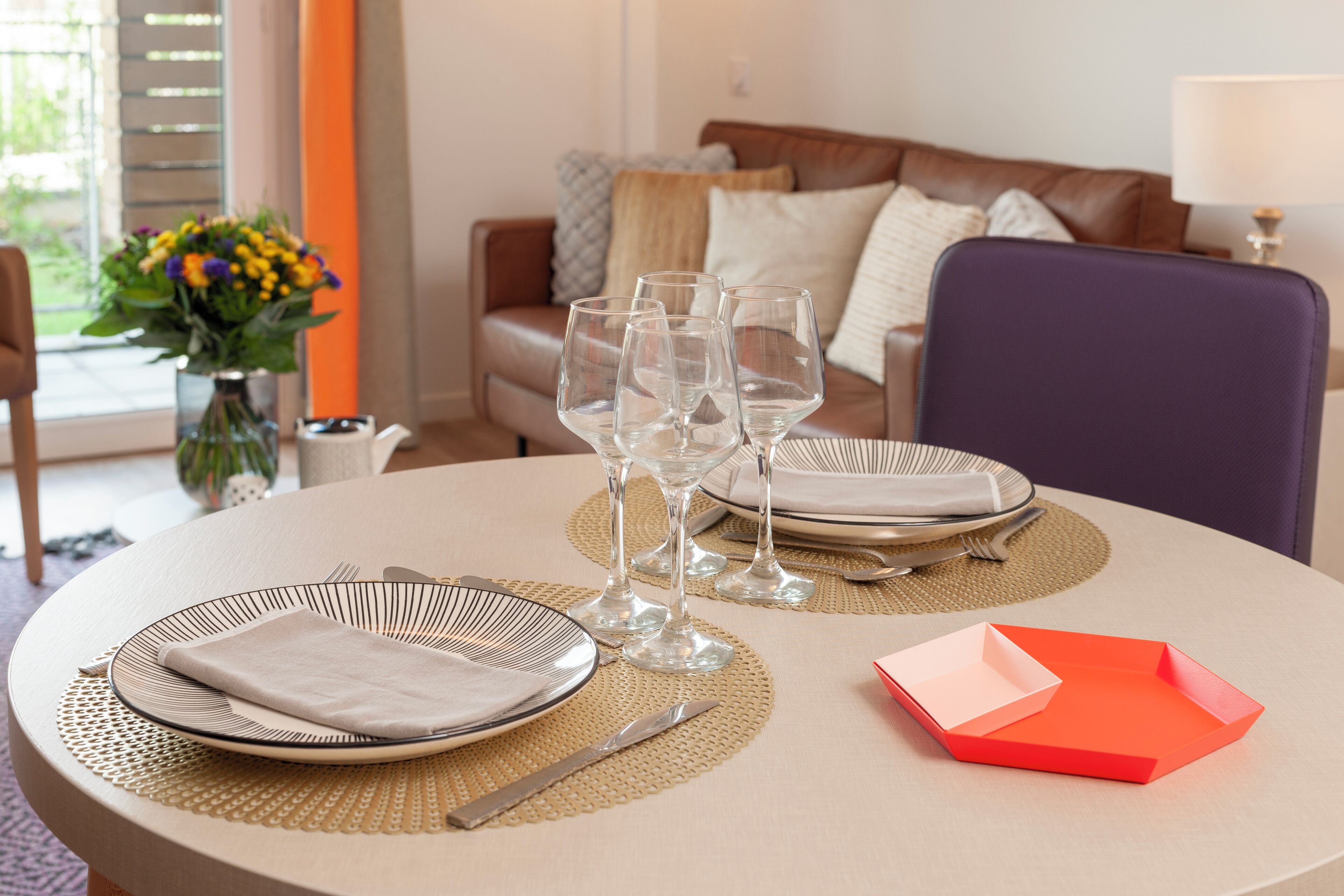 Een eettafel gedekt met borden, bestek en wijnglazen, met een bloemenvaas en een bank op de achtergrond.
