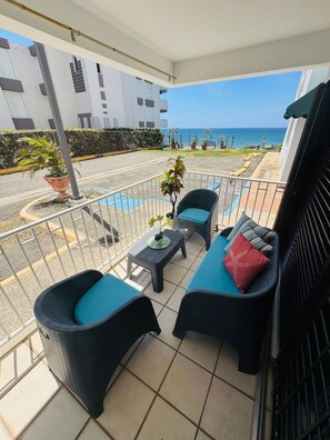 A balcony with two wicker chairs and a small table, overlooking a street and the ocean. - Serenity Sanctuary | Walk to Beach Retreat (Rincón)