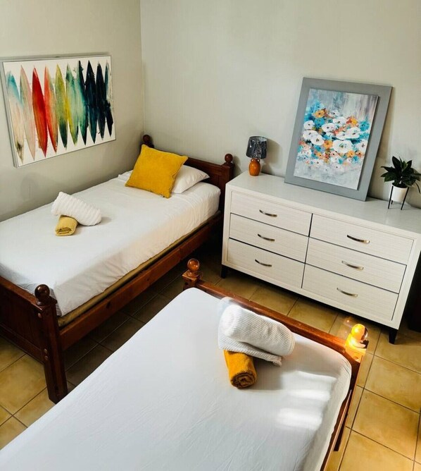 A hotel room with a double bed, a single bed, a bedside table with a lamp, a small cabinet with drawers, and two wall-mounted paintings.