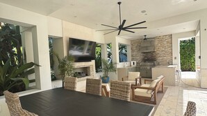 A modern outdoor kitchen with a dining area, including a large dark table and wicker chairs, surrounded by potted plants and a stone backsplash.