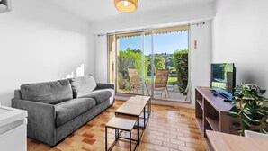 A modern living room with a grey sofa, a wooden coffee table, and a TV on a wooden stand. There is a sliding glass door leading to an outdoor area with chairs and a view of greenery. - Apartment with view on the golf of Houlgate (Houlgate)