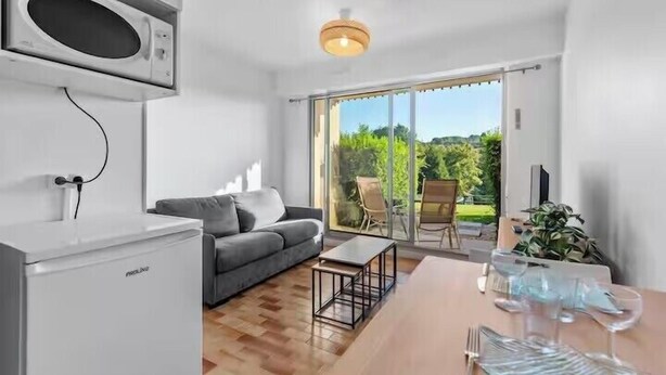 A modern kitchen with a built-in microwave, a grey sofa, a wooden dining table set for two, and a sliding glass door leading to a patio with outdoor seating. - Apartment with view on the golf of Houlgate (Houlgate)