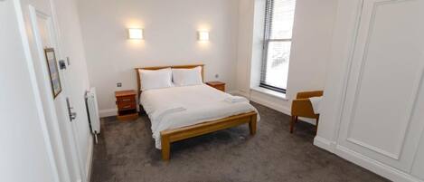 A bedroom with a wooden bed, two bedside tables, a nightstand, a window with blinds, and a door.