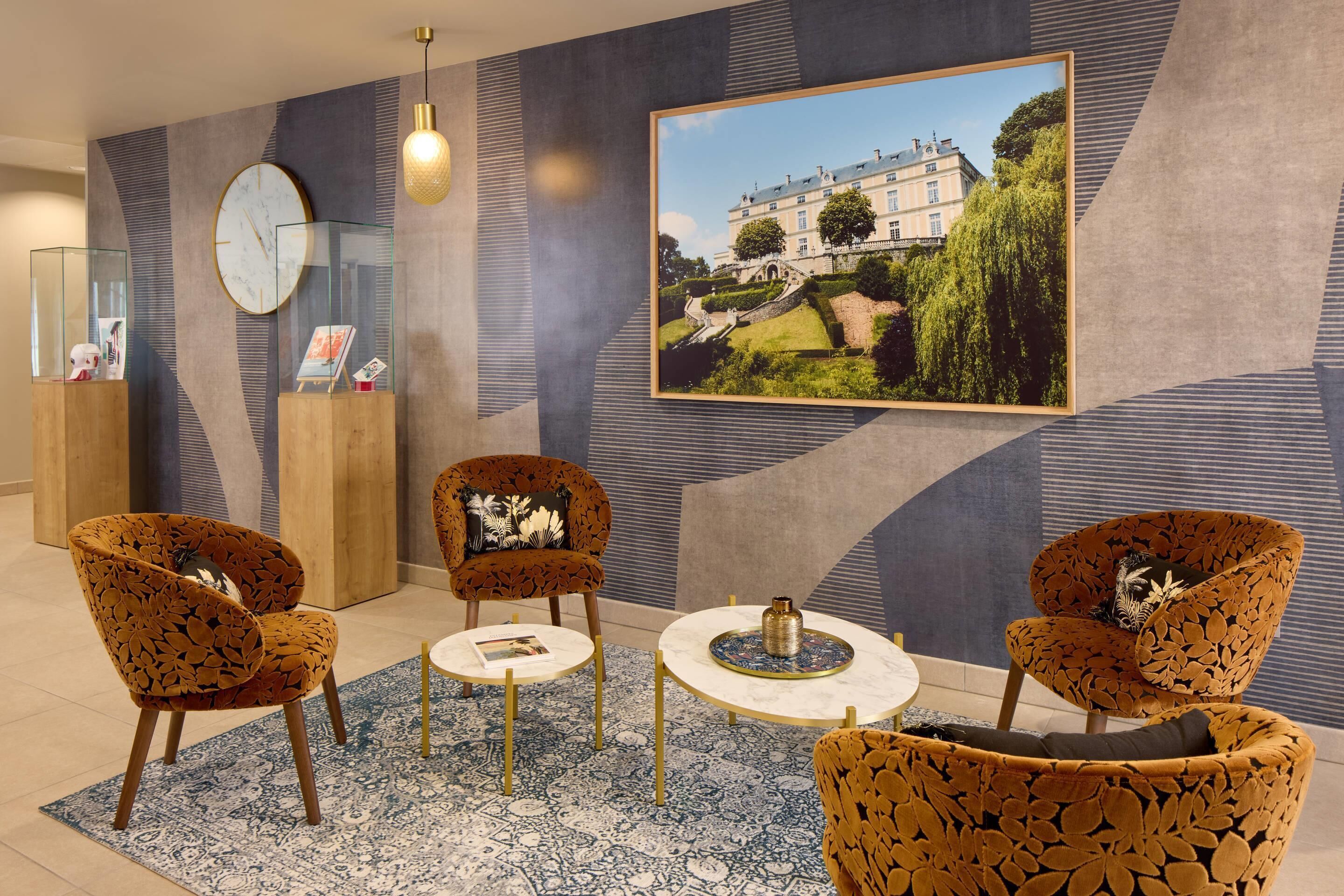 A modern hotel lobby with patterned armchairs, a round table, a clock, and a framed picture of a building.