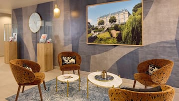 A modern hotel lobby with patterned armchairs, a round table, a clock, and a framed picture of a building.