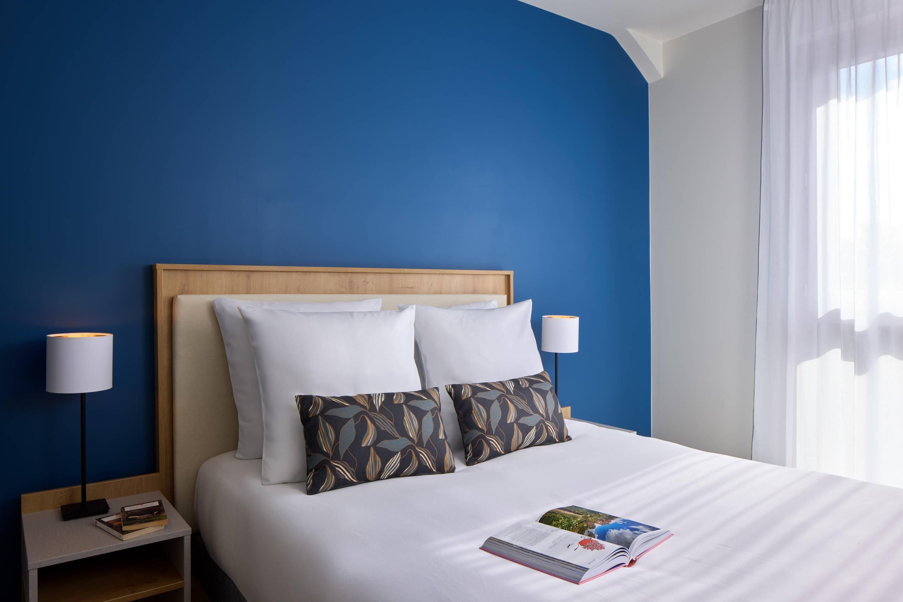 Una cama bien hecha con sábanas blancas y dos almohadones decorativos, una lámpara de noche y un libro sobre la mesita.