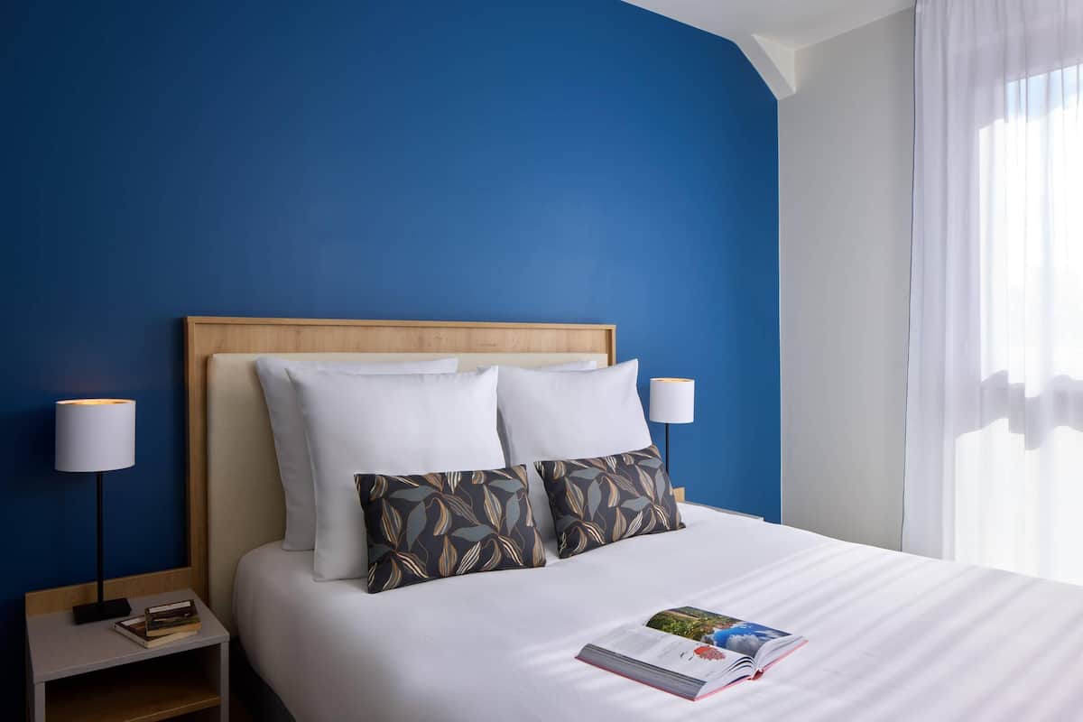 Una cama bien hecha con sábanas blancas y dos almohadones decorativos, una lámpara de noche y un libro sobre la mesita.