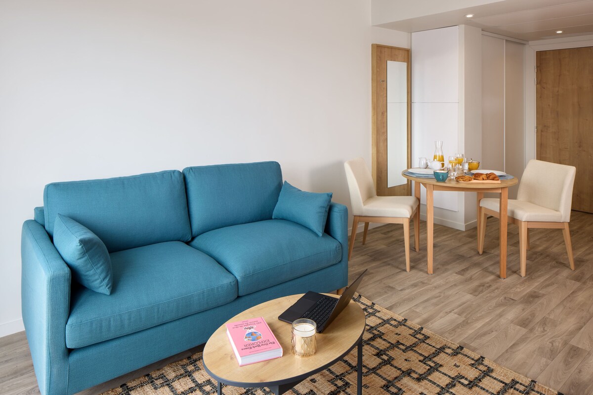 A living room with a blue sofa, a small round table with a laptop, a book, and a glass of milk.