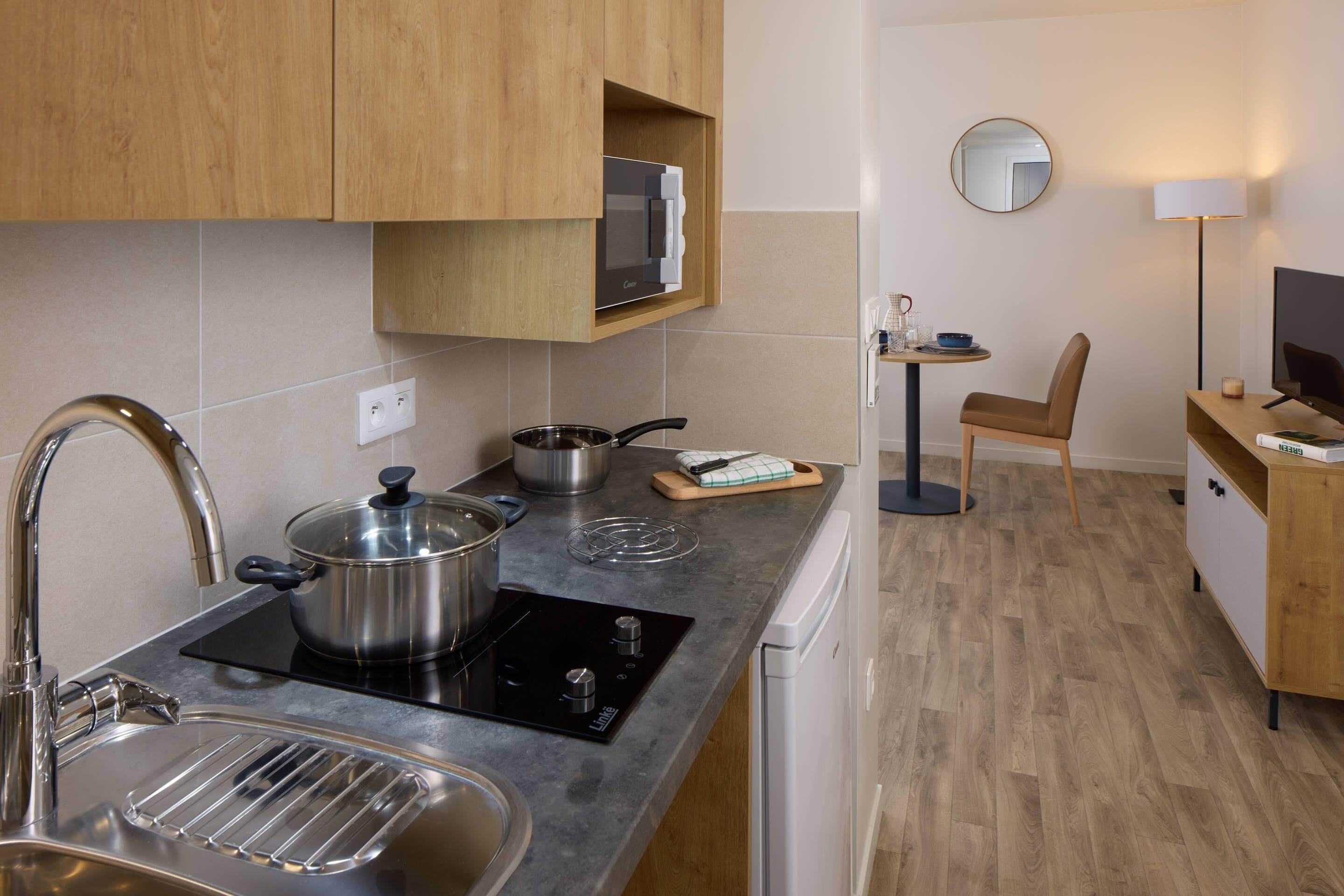 Une cuisine compacte équipée d’un évier, d’une cuisinière et d’un four à micro-ondes. On y trouve une chaise et une petite table avec une lampe.