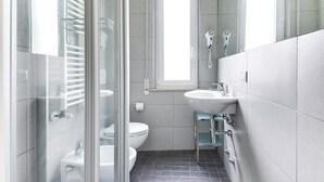 A modern bathroom with a glass shower enclosure, a white toilet, a sink with a mirror, and a window providing natural light. - Three-room apartment Scola 11 (Gabicce Mare)