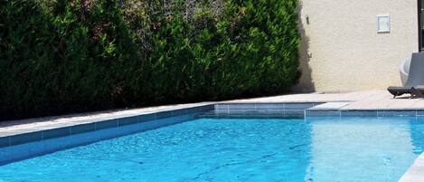 Ein Schwimmbecken mit einer Fliesenverkleidung, umgeben von einer Hecke und einem Gebäude mit einer hellen Wand.