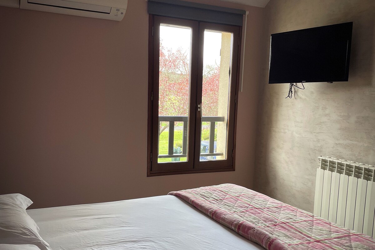 Um quarto com cama, janela, radiador e televisão fixada na parede.