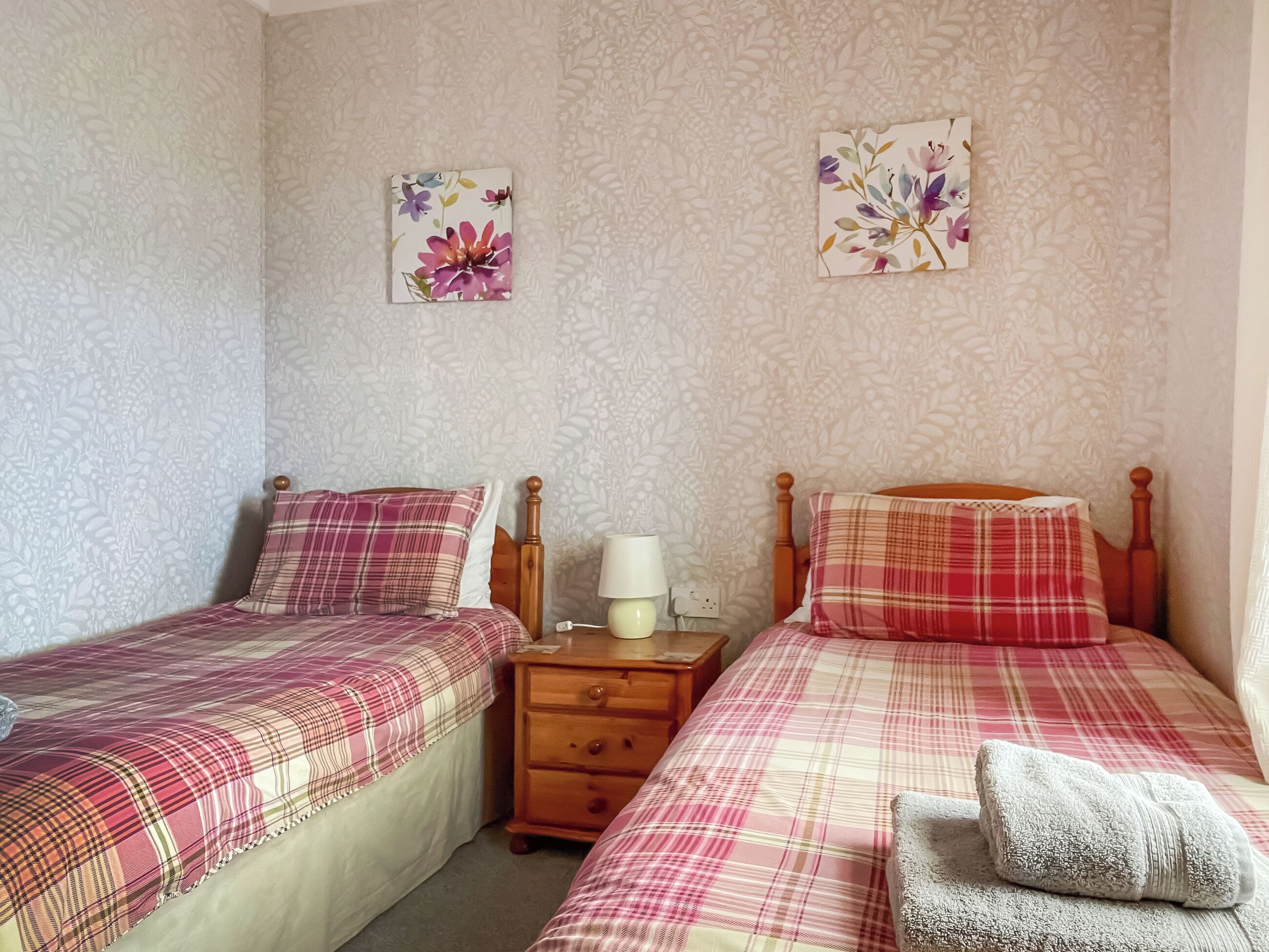 A bedroom with two beds, each with a plaid bedspread, a wooden nightstand with a lamp, and floral artwork on the wall.