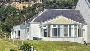 Ein weißes Gebäude mit Erker, einer kleinen Veranda und einem grasbewachsenen Hügel im Hintergrund.