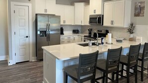 A modern kitchen with white cabinetry, a stainless steel refrigerator, a microwave, and a large island with bar stools. - Luxury Condo at Crow Creek golf resort (Brunswick County)