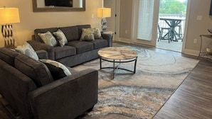 A living room with a sofa set, a coffee table, and a large rug. There is a door leading to a balcony with outdoor furniture. - Luxury Condo at Crow Creek golf resort (Brunswick County)