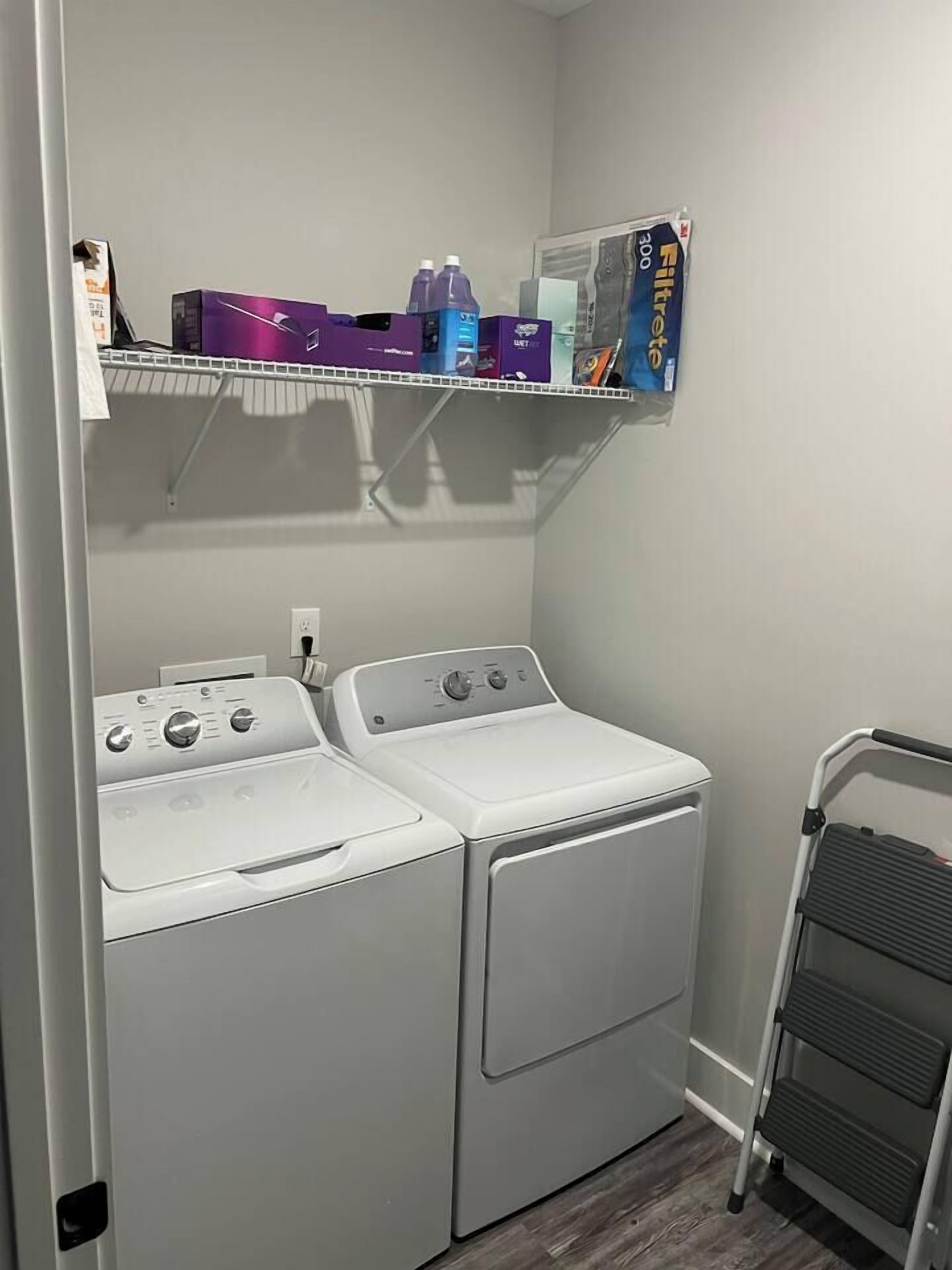 A laundry room with two white front-loading washers and dryers, a metal step ladder, and shelves above holding various laundry supplies.
