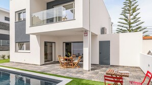 Een modern huis met twee verdiepingen, een zwembad, buitenmeubilair en een zichtbaar alarmsysteem.