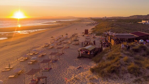 Een strand met rijen ligbedden en parasols, een klein kioskje en een gebouw met een rood dak op de achtergrond.