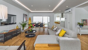 Een moderne woonkamer met een grijze bank, een salontafel en een televisie. Er is een eethoek met stoelen en een tafel, en een keuken met een witte kast en een plant.