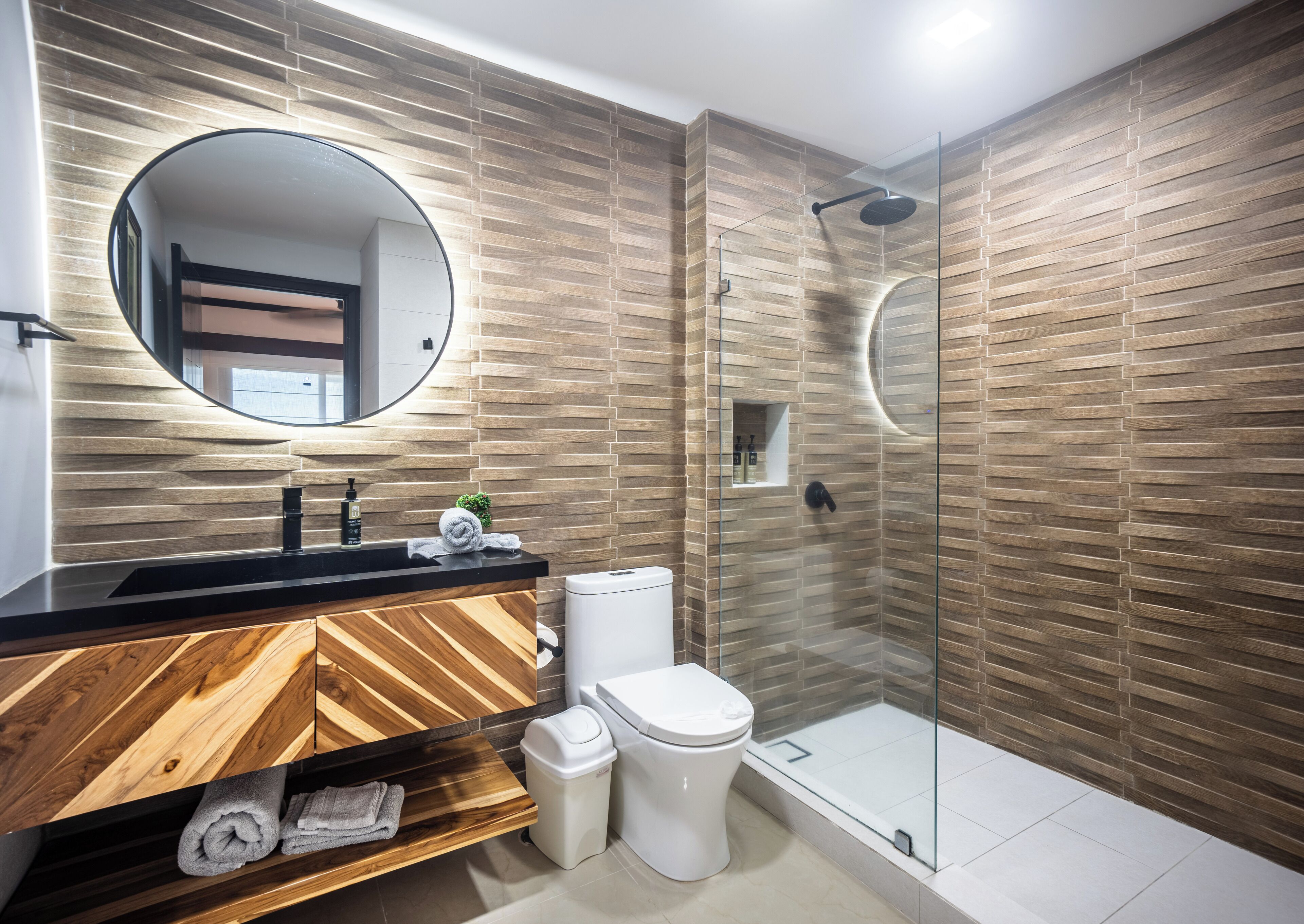 Ein modernes Badezimmer mit einem Holzbidet, einem runden Spiegel, einer Toilette und einer Duschkabine.