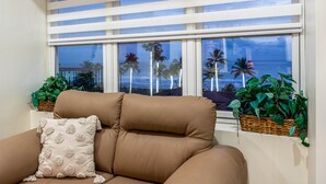 Un sofá color arena con un cojín decorativo, una ventana con persianas a rayas y dos cestas de mimbre con plantas.