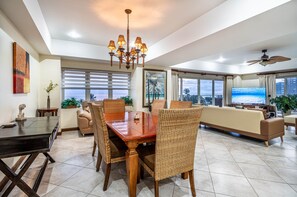 A spacious living area with a dining table, wicker chairs, a sofa, and a large TV screen.
