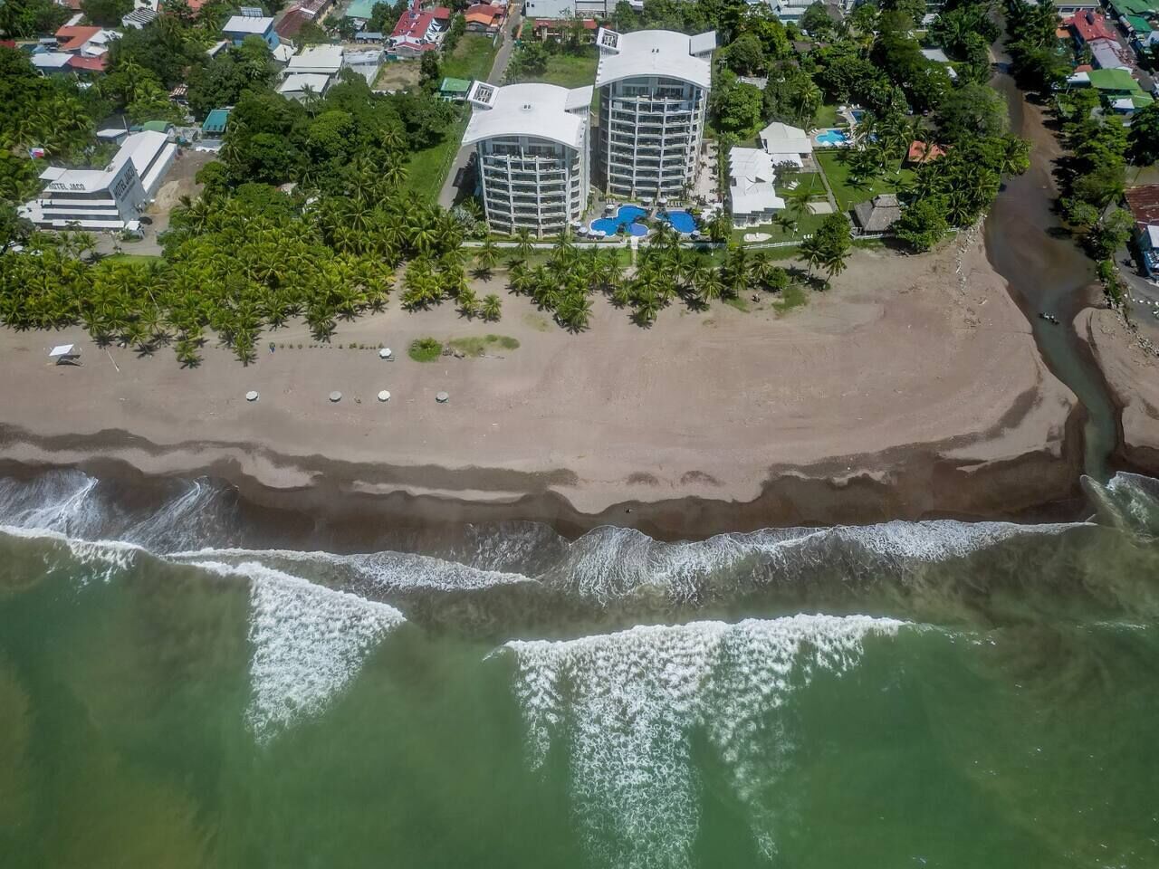Eine Luftaufnahme einer Küstenregion mit Strand, zwei großen Gebäuden mit Balkonen und einem Schwimmbad.
