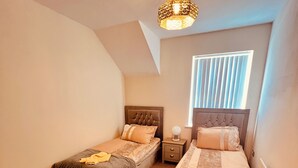 A bedroom with two beds, a nightstand, a lamp, and a window with blinds. - Relaxing Enjoyable Stay IN middlesbrough TS5 (Middlesbrough)