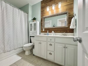 A bathroom with a shower curtain, toilet, vanity with a mirror, and a sink. - The Vida Sol! Steps to the Ocean. Sound views, Sunsets, Pier + Kayak launch (North Topsail Beach)