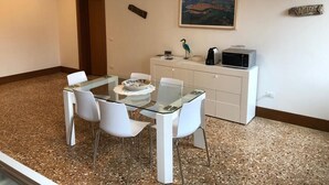 Un comedor con mesa de vidrio y sillas blancas, una cocina con microondas, y un cuadro de paisaje en la pared.