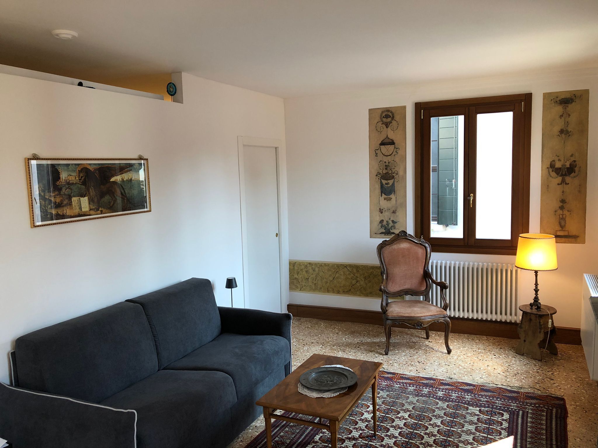 A living room with a dark sofa, a wooden coffee table, a patterned rug, a chair, a radiator, and wall art.